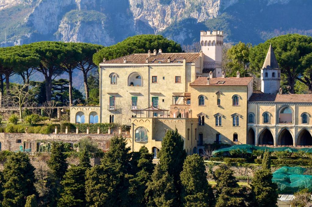 A hillside villa of pale stone and red roofs, with pines and mountains behind it.