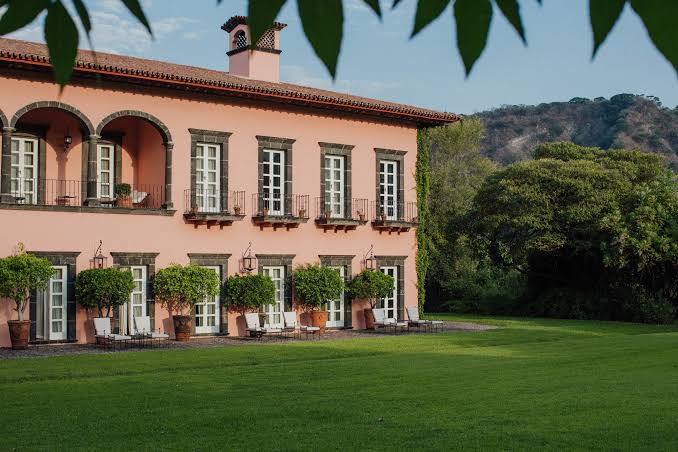 A pink hacienda with a lawn, terrace chairs and hills beyond.