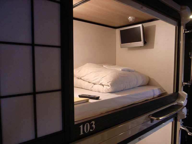 The inside of a compact sleeping capsule with white bedding and a small screen.