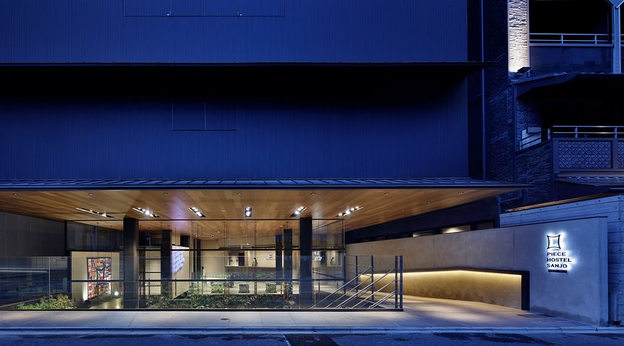 The night entrance of Piece Hostel Sanjo, with a wooden canopy and glass doors.